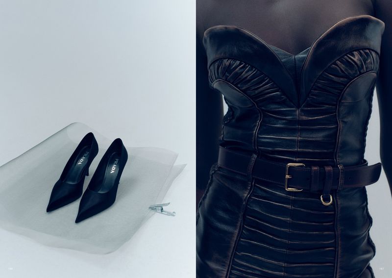 Fashion still life featuring a pair of black Prada heels and a black leather dress with a brown belt against a white backdrop.