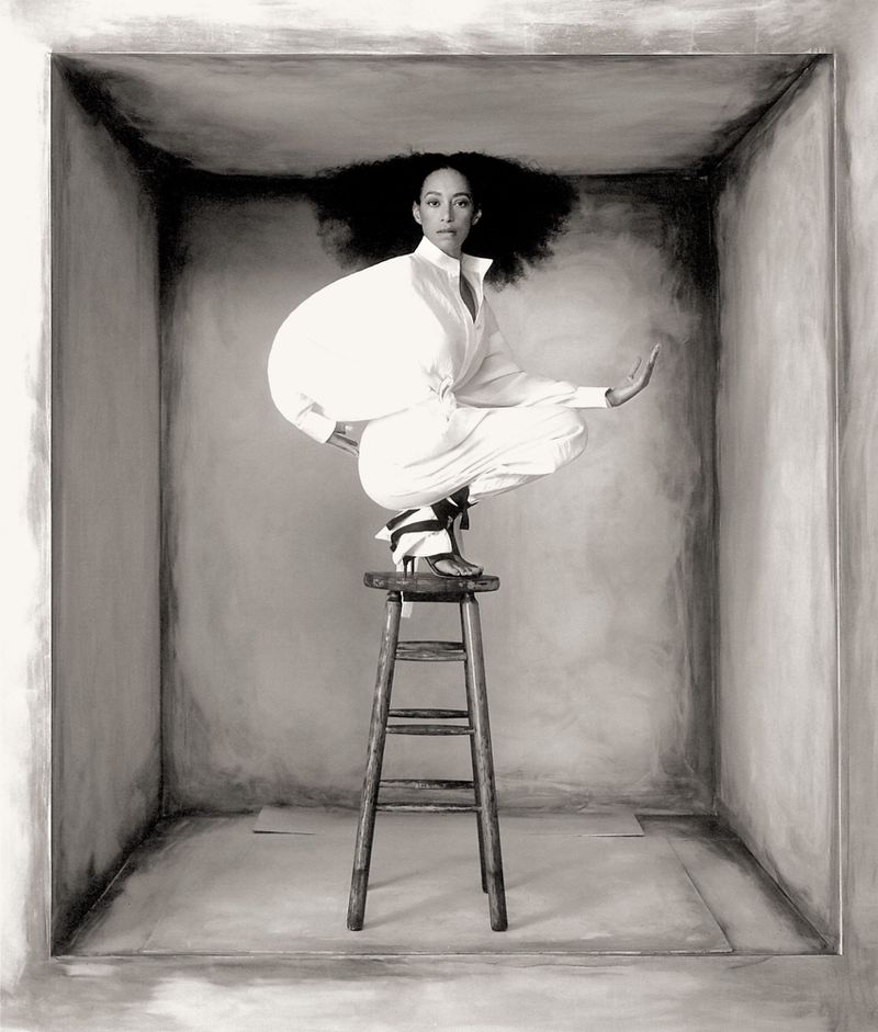 Black and white studio shot of a stylish adult woman in a white outfit and black heels, posed on a wooden stool in a grey box.