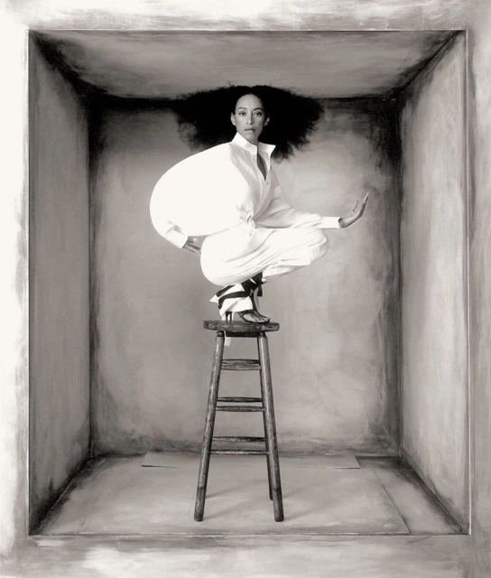 Black and white studio shot of a stylish adult woman in a white outfit and black heels, posed on a wooden stool in a grey box.