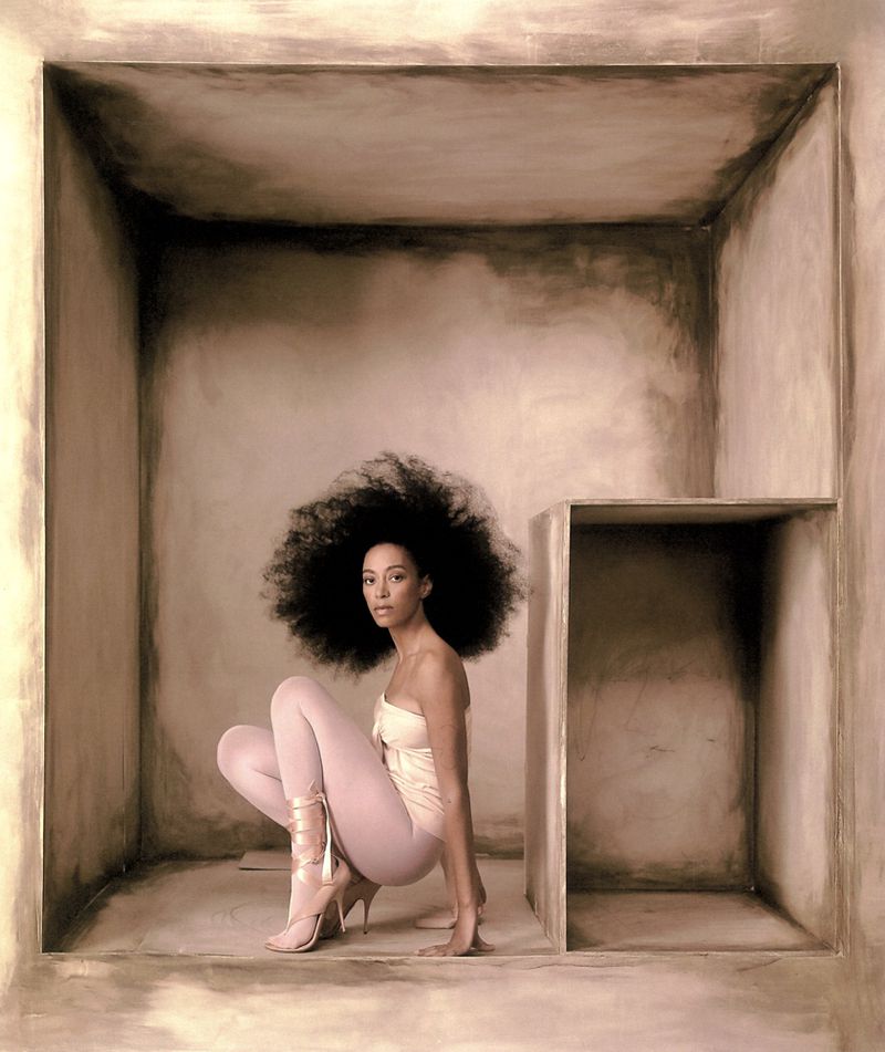 Woman with large afro in strapless top, leggings, and heels crouching inside of square structure in a photo studio setting.