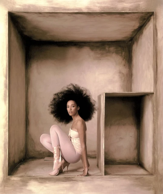 Woman with large afro in strapless top, leggings, and heels crouching inside of square structure in a photo studio setting.