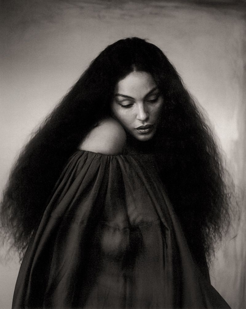 Monochrome portrait of a woman with long, dark hair, wearing a draped garment and gazing downward in a studio setting with soft lighting.