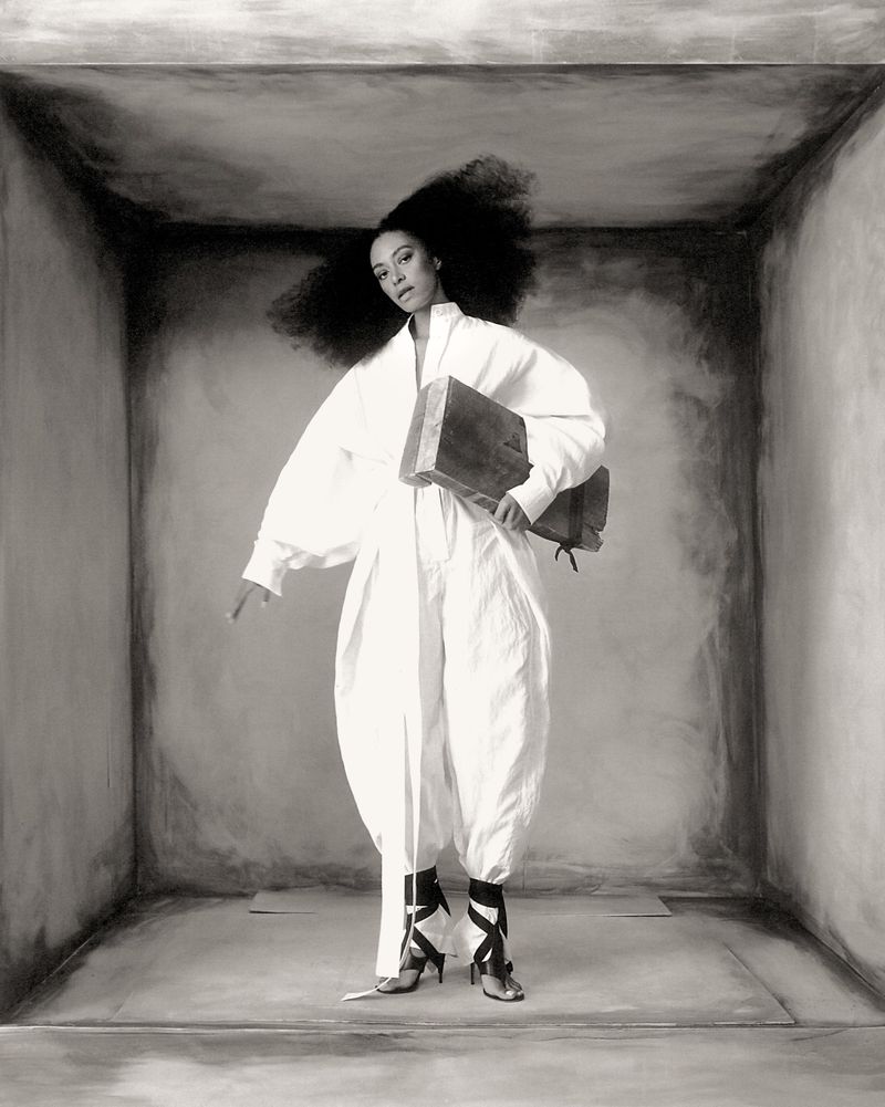 Black and white portrait of a woman in a white jumpsuit with dark hair, holding a dark book, standing in a white box.