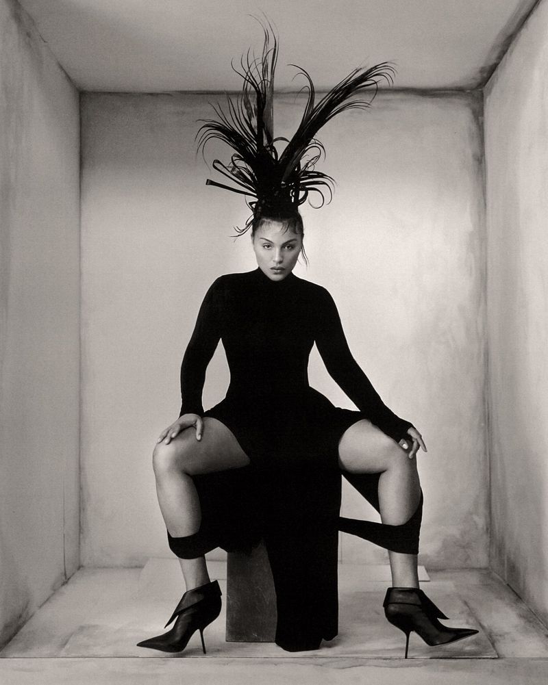 Monochrome portrait of a woman in a black dress and high heels, seated on a block inside a bare room with high-fashion hairstyling.