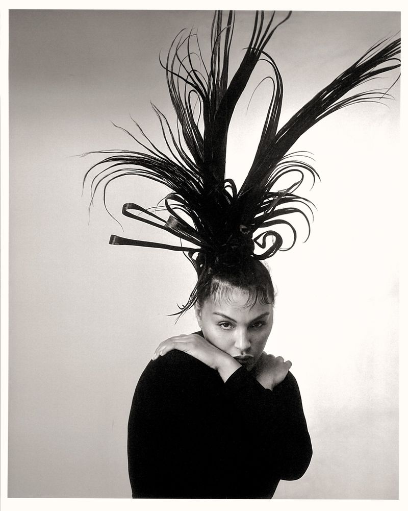 Monochrome portrait of a person with dramatic dark hair art, wearing a black top, standing in a light studio setting.