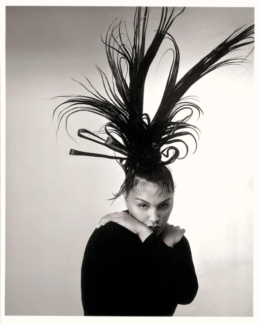 Monochrome portrait of a person with dramatic dark hair art, wearing a black top, standing in a light studio setting.