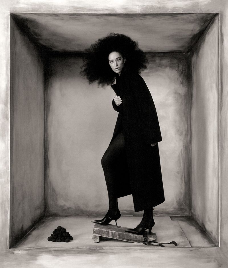 Monochrome fashion shot of a woman in a dark coat and heels stepping on a wooden block inside a painted square, artistic expression.