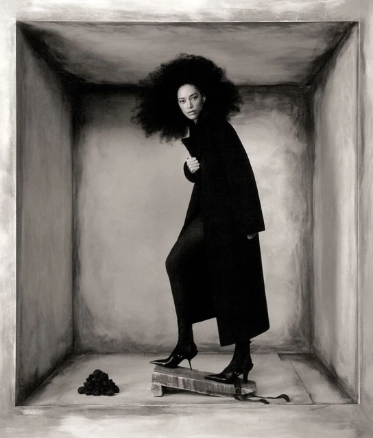 Monochrome fashion shot of a woman in a dark coat and heels stepping on a wooden block inside a painted square, artistic expression.