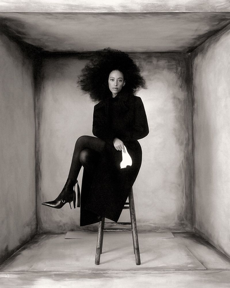 Monochrome portrait of a Black woman with a large afro sitting on a stool in a gray box, wearing a dark coat, boots, and holding a handkerchief.