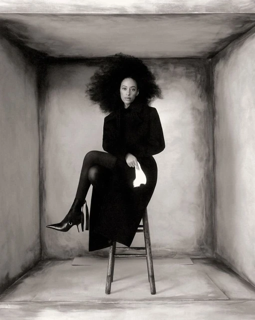 Monochrome portrait of a Black woman with a large afro sitting on a stool in a gray box, wearing a dark coat, boots, and holding a handkerchief.