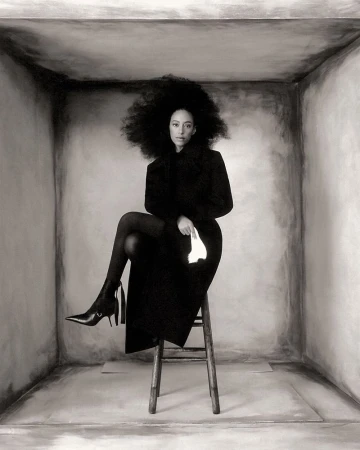 Monochrome portrait of a Black woman with a large afro sitting on a stool in a gray box, wearing a dark coat, boots, and holding a handkerchief.