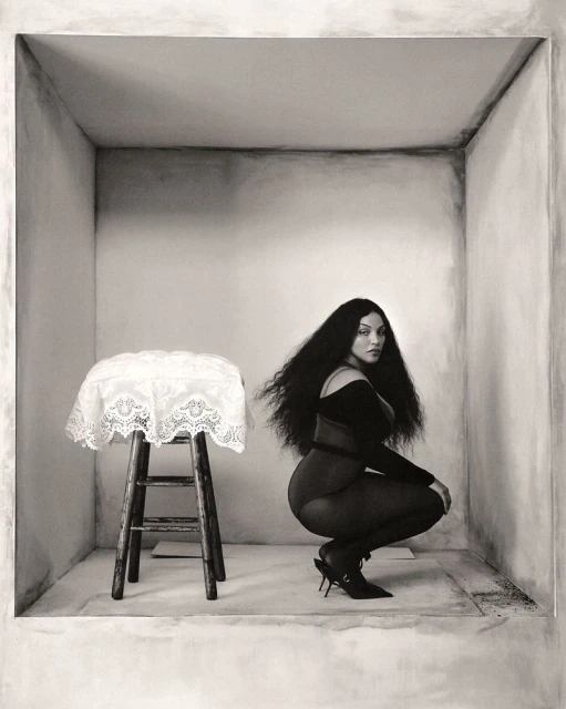 Monochrome portrait of a woman with long dark hair, crouching near a stool with a lace cover, inside of a minimally decorated square space.