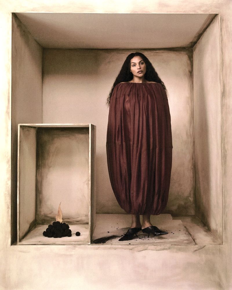 Adult woman wearing an oversized brown dress and black heels standing inside a white box, with an art installation to the left.