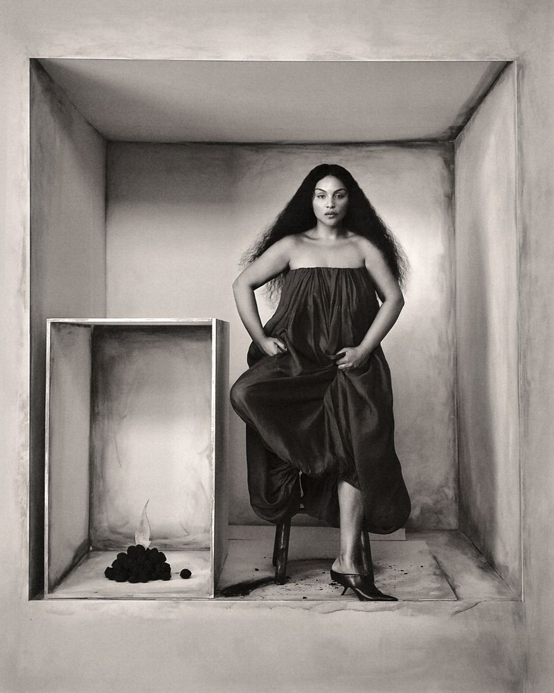 Elegant adult female posing on a stool inside a painted box, wearing a strapless dress and heels, next to a pile of objects.  