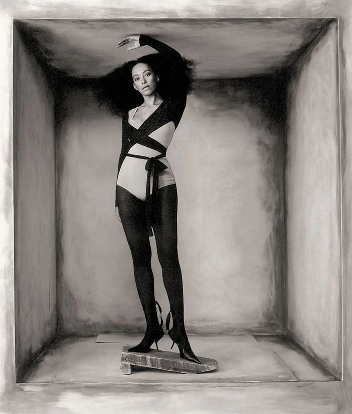 Model Paloma Elsesser posing inside a textured rectangular box set wearing a strappy bodysuit and dark tights