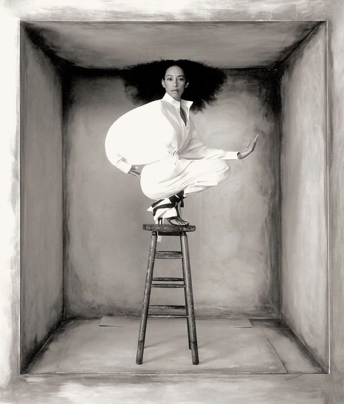 Model Paloma Elsesser wearing white clothing squatting on a wooden stool inside a large box-like set for Harper's Bazaar Italia