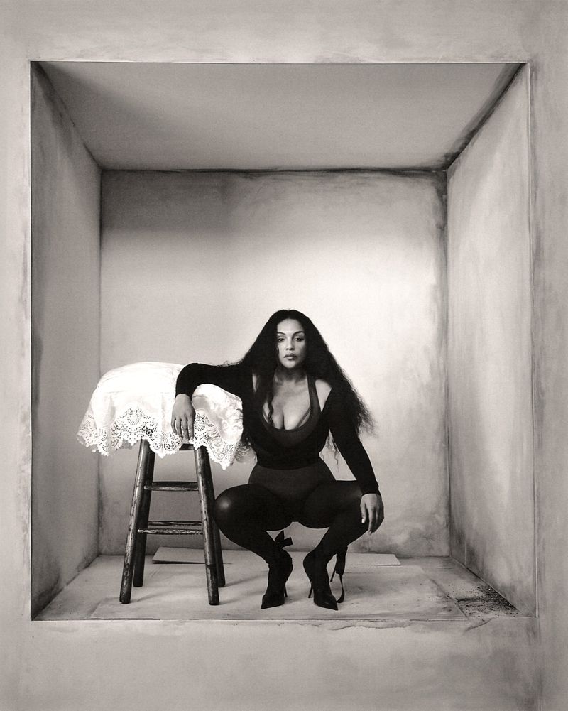 Paloma Elsesser crouching inside a structured grey alcove beside a small wooden stool draped with a white lace cloth.