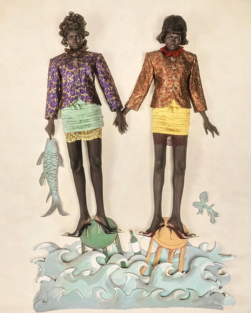 Two mannequins in patterned jackets and mini skirts stand on stools above painted waves, one holding a fish sculpture.