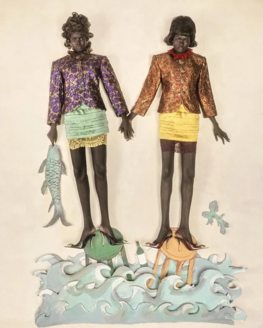 Two mannequins in patterned jackets and mini skirts stand on stools above painted waves, one holding a fish sculpture.