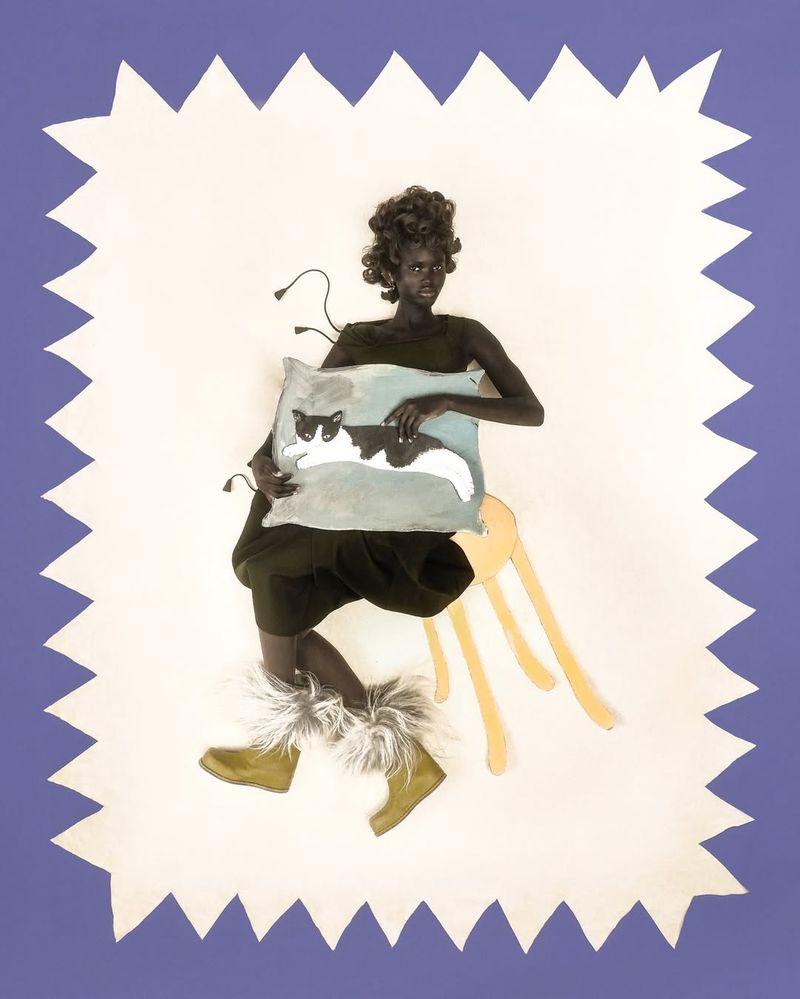 A woman with an afro wearing fur boots and a black dress, sits on a yellow stool and holds a pillow with a cat print in a studio.