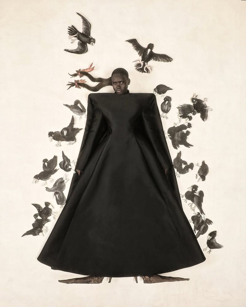 An African woman wearing a long black dress surrounded by black-and-white bird illustrations creates a surreal fashion portrait.
