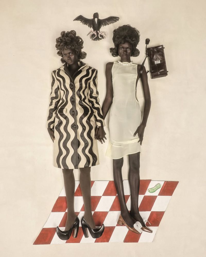 Two women in vintage-inspired fashion pose with a bird figure and purse, standing on a red and white checkered surface against a white backdrop.