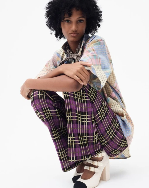 A fashion shot presents a female model with dark curly hair in a patchwork top and purple plaid pants, kneeling against a plain white backdrop.