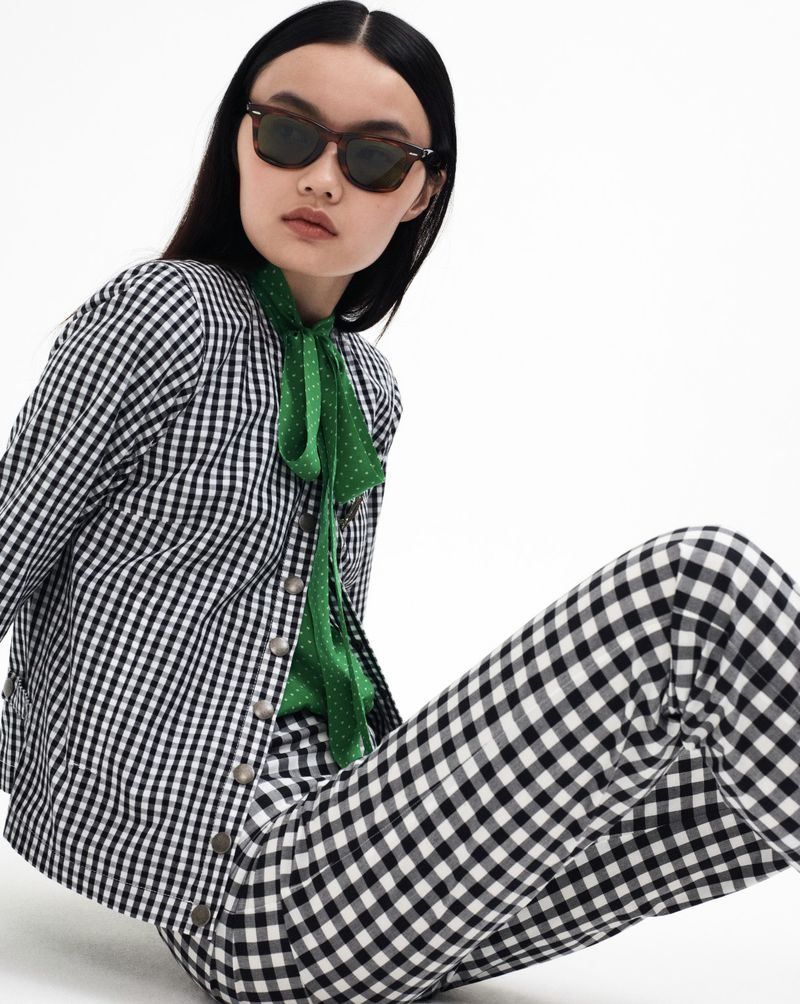 Brunette woman poses in black and white gingham jacket, green polka dot blouse, and matching pants against a bright white backdrop.