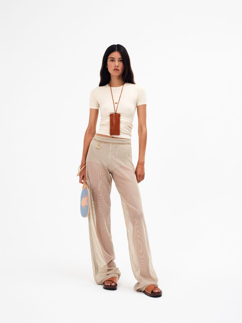 Full-length studio shot of a woman wearing a white top and patterned semi-sheer trousers, accessorized with brown sandals and a cellphone case.