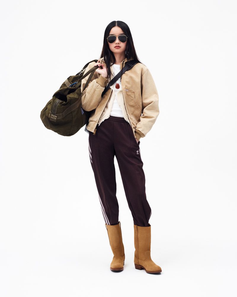 Adult female in beige jacket and track pants with olive duffel bag against a white backdrop in a full-length studio shot.