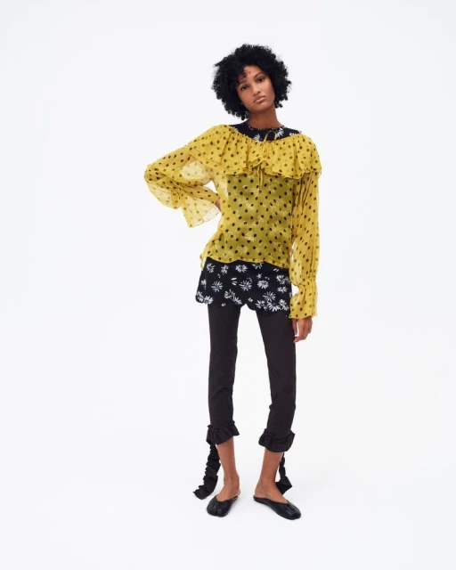 An African-American fashion model in a yellow polka dot blouse, black floral mini-skirt, and leggings poses in a white photo studio.