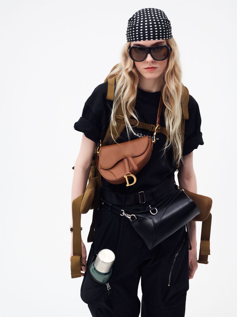 A young adult woman with long blonde hair wears a black t-shirt, black pants, and multiple bags, including a Dior saddlebag.