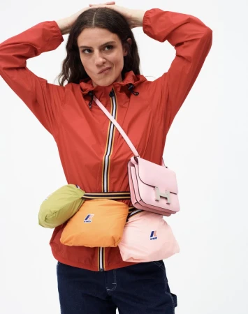 Woman in red hooded jacket with colorful waist pack and pink purse against white background in a studio lit setting.