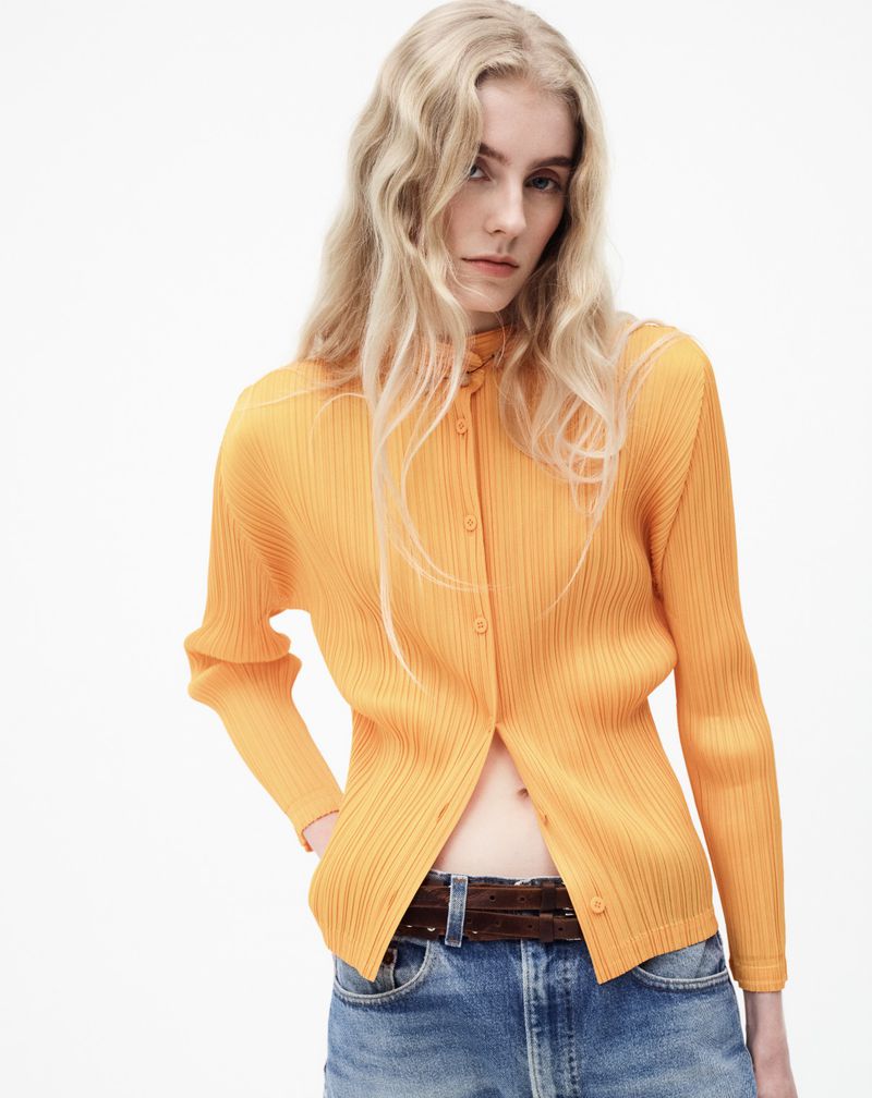 A woman with blonde hair is wearing an orange ribbed cardigan and blue jeans against a white backdrop in a fashion-style studio shot.