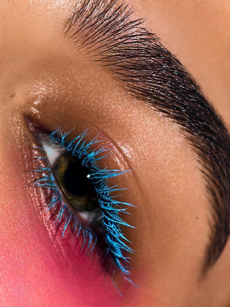 Extreme close-up on a woman's eye with long, bright-blue mascara on the eyelashes, dark eyebrow and pink blush on the cheekbone.