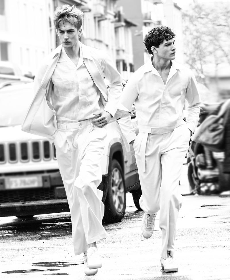 Two young adult men in all-white outfits walk on a city street near a car. Black and white street style fashion editorial.