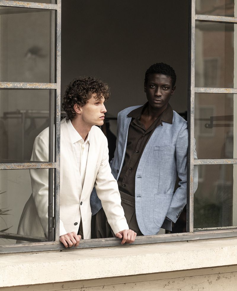 Two men in jackets pose in a window frame, one with curly hair in a cream jacket, the other with dark skin in a blue jacket.