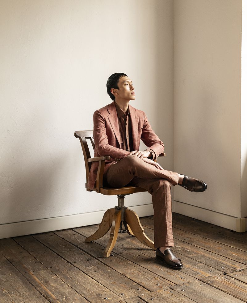 An Asian man in a brown suit sits with legs crossed on a vintage wooden swivel chair against a neutral wall in a brightly lit room.