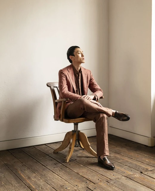 An Asian man in a brown suit sits with legs crossed on a vintage wooden swivel chair against a neutral wall in a brightly lit room.