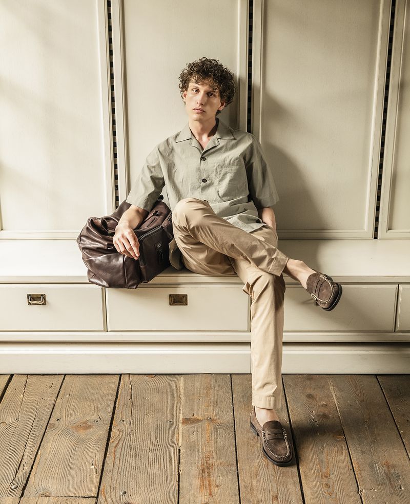 A young man sits on a vintage cabinet wearing a green, short-sleeved shirt and khaki pants, complemented by a brown leather duffel bag.