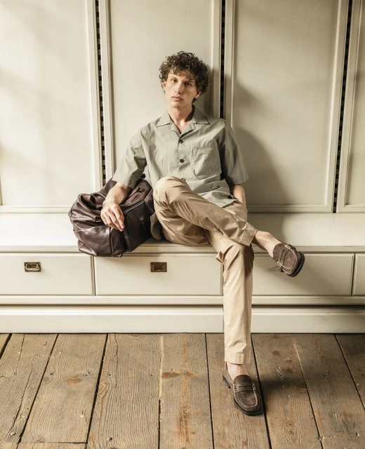 A young man sits on a vintage cabinet wearing a green, short-sleeved shirt and khaki pants, complemented by a brown leather duffel bag.