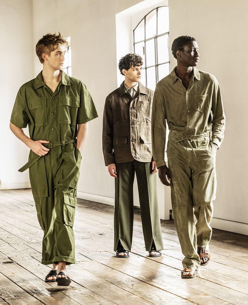 Three male models wearing olive green and brown outfits walk on a wooden floor in a bright indoor fashion shoot with white walls.