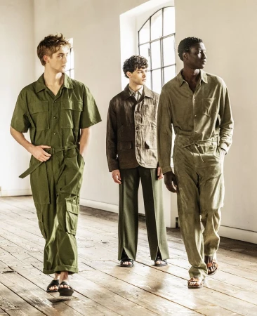 Three male models wearing olive green and brown outfits walk on a wooden floor in a bright indoor fashion shoot with white walls.