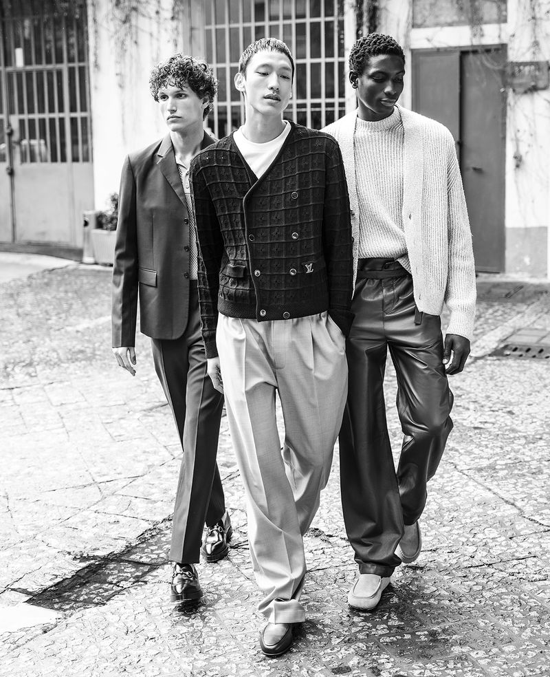 Three male fashion models walk outdoors on a city street in monochrome, wearing stylish suits and sweaters with a blurred background.