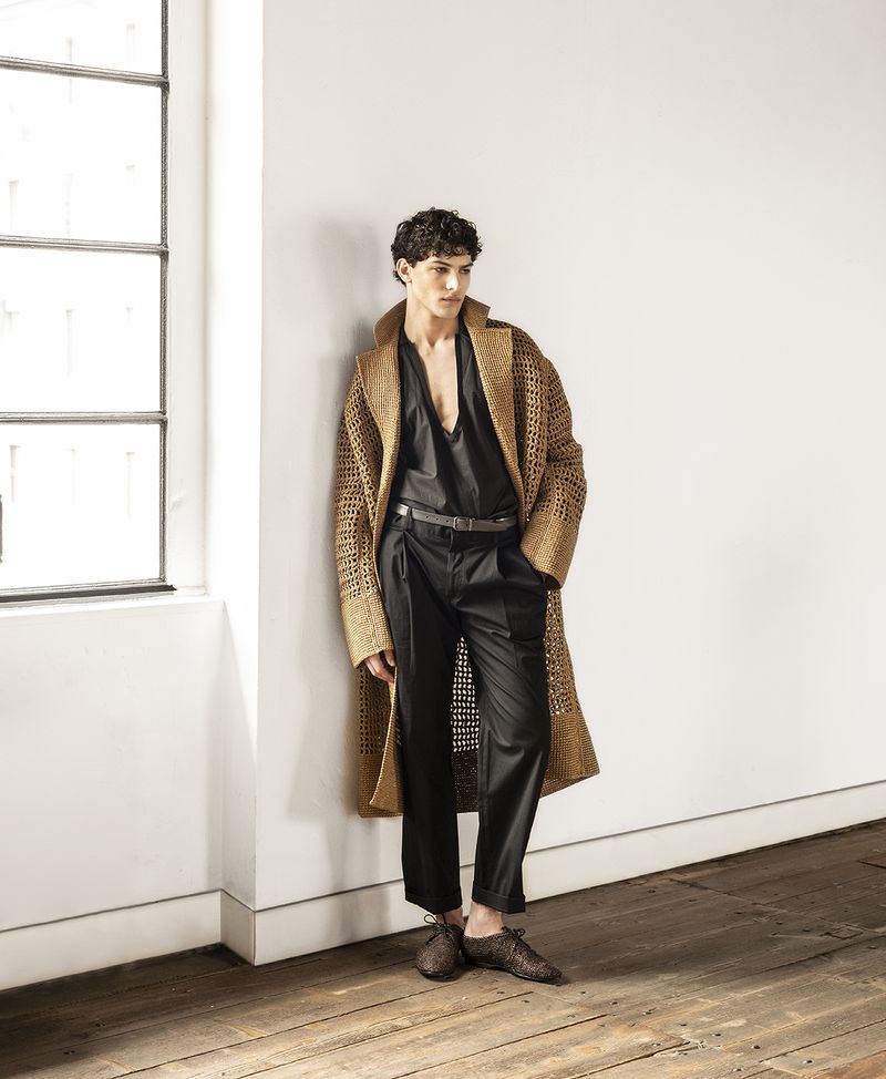 Fashionable man in unbuttoned black top and black pants posing against a white wall with a window, wearing brown coat and loafers.
