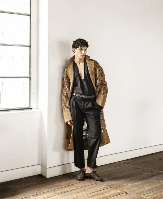 Fashionable man in unbuttoned black top and black pants posing against a white wall with a window, wearing brown coat and loafers.