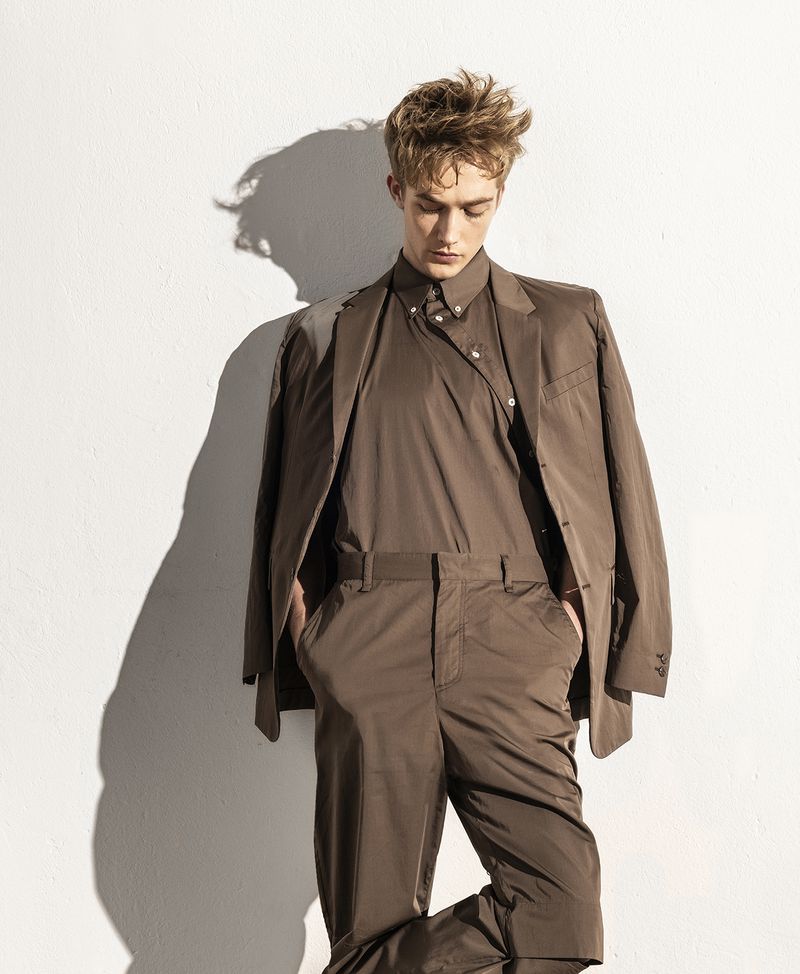 Portrait of a young adult man in a brown suit, posing against a white wall with shadows. Fashion shot with studio lighting.