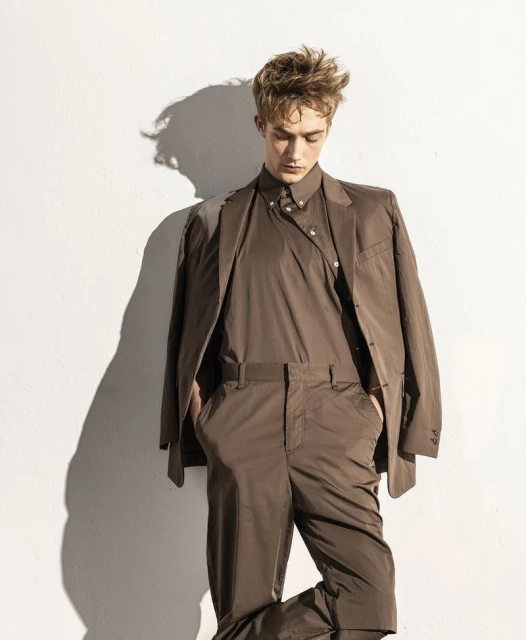 Portrait of a young adult man in a brown suit, posing against a white wall with shadows. Fashion shot with studio lighting.