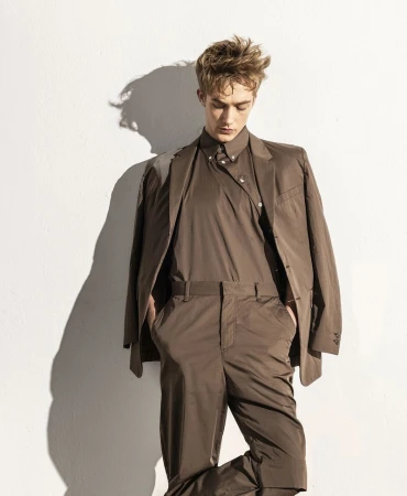 Portrait of a young adult man in a brown suit, posing against a white wall with shadows. Fashion shot with studio lighting.