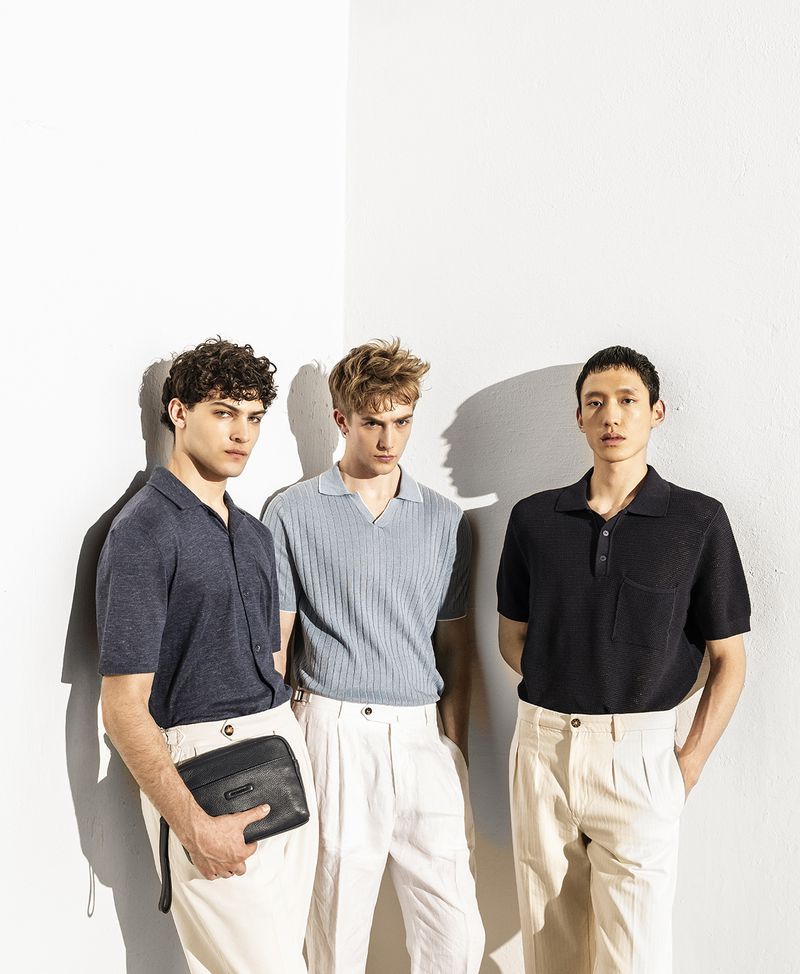 Three adult men stand in a photo studio wearing polo shirts and light-colored pants. One man holds a black clutch. Studio lighting.